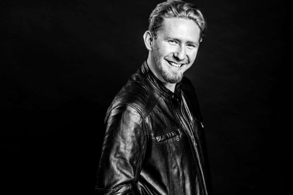 A headshot of Galen White; a blonde, Caucasian man in smiling wearing a leather jacket and smiling at the camera.